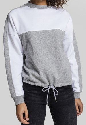 Frau trägt einen grau-weißen Sweatshirt mit langen Ärmeln und Kordelzug am Saum sowie schwarze Hosen, steht vor einem einfarbigen Hintergrund.