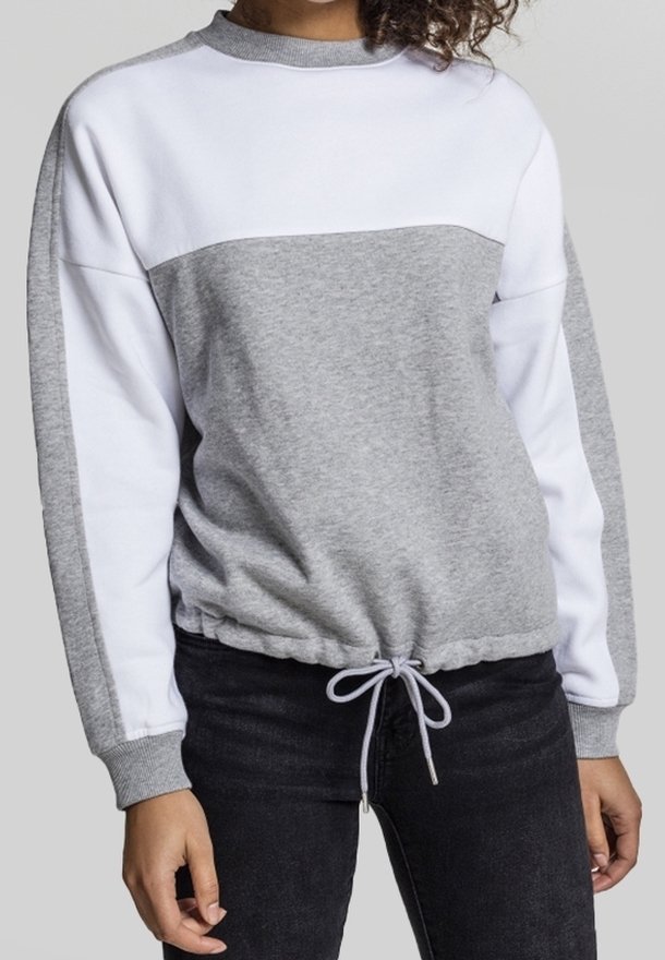 Femme portant un sweat-shirt gris et blanc à manches longues avec cordon de serrage à l'ourlet et pantalon noir, debout devant un fond uni.