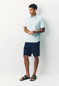 Camicia azzurro chiaro a maniche corte con piccoli motivi di palme, abbinata a pantaloncini blu navy e sandali neri. Tessuto liscio con vestibilità comoda.