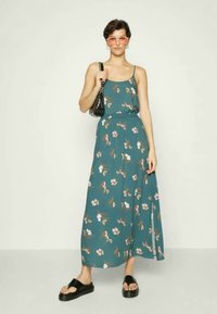 Vestido maxi floral em teal com alças finas, cintura ajustada, fenda lateral e tecido fluido; combinado com sandálias plataforma pretas e uma bolsa.