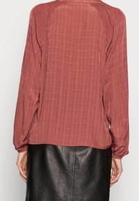 Blusa di colore ruggine con maniche lunghe, tessuto testurizzato con sottili righe, indossata sopra una gonna in pelle nera.