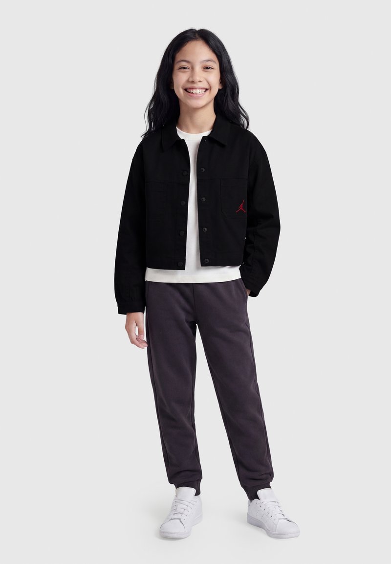 Fille souriante aux longs cheveux foncés porte une veste noire par-dessus une chemise blanche, un pantalon foncé et des baskets blanches, debout devant un fond uni.