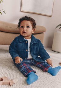 Chaqueta de pana azul con dos bolsillos frontales, camisa blanca, pantalones de cuadros en azul y rojo, y calcetines azules. Sentado sobre una alfombra texturizada.