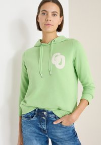 Vrouw die een lichtgroene hoodie met een wit logo en een blauwe spijkerbroek draagt, staand met één hand in de zak tegen een effen achtergrond.