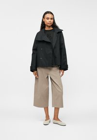 Abrigo trinchera negro de doble botonadura con solapas anchas, combinado con una camisa negra con un logo en relieve y pantalones de pierna ancha beige.