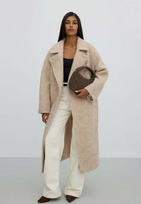 Žena s dlouhými tmavými vlasy, oblečená v béžovém dlouhém kabátu, černém topu, bílých kalhotách a hnědých špičatých botách, držící hnědou kabelku na jednobarevném pozadí.