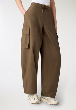 Pantaloni cargo marroni a gamba larga con tasche laterali a patta, indossati con sneakers bianche e top bianco corto.