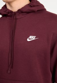 Vínová mikina Nike s kapucí na zavazování, bílým logem na hrudi, vyrobená z měkké látky s hladkým povrchem.