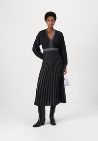 Musta poimutettu mekko V-pääntie-designilla, pystysuorat hopeasuoristukset, pitkät hihat ja tiukka vyötärö. Yhdistetty polvipituisille mustille saapaille ja valkoiselle laukulle.