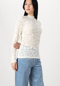 Haut à manches longues en dentelle couleur blanc cassé avec motif floral ; design à col montant ; porté avec un pantalon en velours côtelé bleu clair.