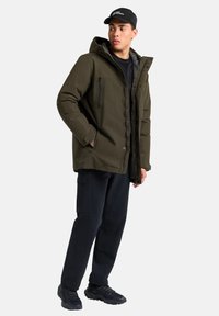 Giacca a vento verde oliva con cappuccio, bottoni frontali e tasche con cerniera. Indossata con pantaloni neri e sneakers nere texturizzate.