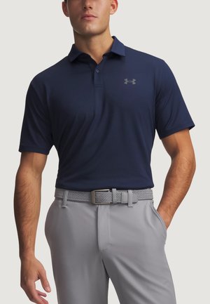 Uomo che indossa una polo a maniche corte blu navy con logo Under Armour, pantaloni grigio chiaro e cintura grigia intrecciata, in piedi con una mano in tasca.
