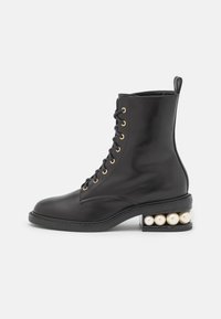 Botín de cuero negro hasta el tobillo con ojales dorados, diseño con cordones y un tacón único que presenta grandes perlas decorativas blancas. Textura suave, forma moderna.