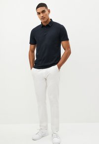 Marineblaues Polohemd mit Kragen und einer Drei-Knopf-Leiste, kombiniert mit weißen Hosen und weißen Sneakern, vor einem neutralen Hintergrund stehend.