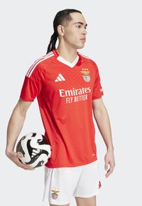 Červený futbalový dres s bielymi akcentmi, s výstrihom do V a diagonálnym vzorom. V kombinácii s bielymi šortkami a čierno-bielym futbalovým loptou.