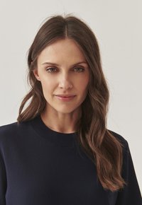 Marineblauwe sweatshirt met een ronde hals en geribbelde textuur, met lange, golvende bruine haren, genomen tegen een lichte achtergrond.