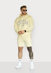 Light yellow hoodie with "ESTEEM" graphic, paired with matching shorts. White sneakers and crew socks, featuring a visible tattoo on the leg.