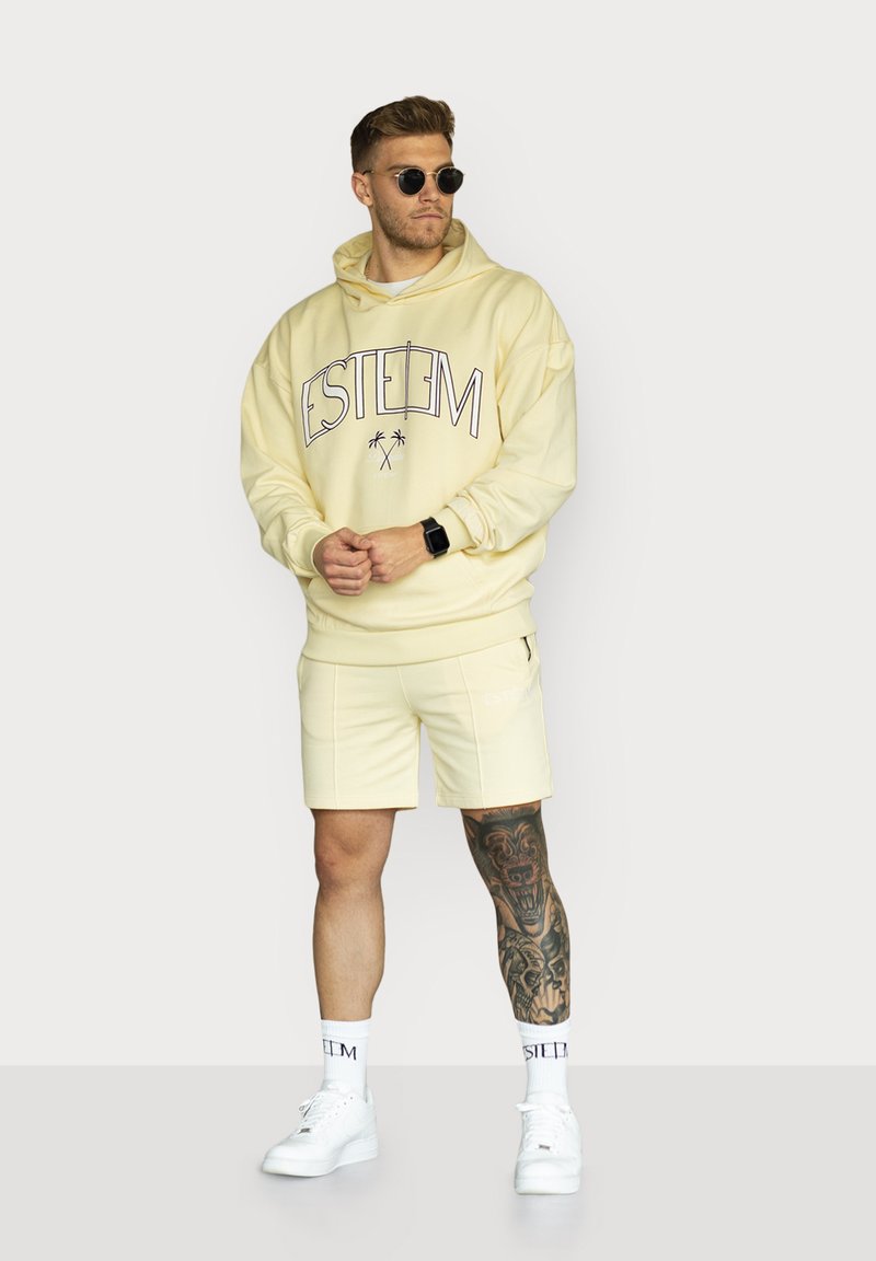 Light yellow hoodie with "ESTEEM" graphic, paired with matching shorts. White sneakers and crew socks, featuring a visible tattoo on the leg.