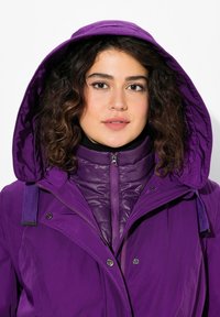 Giacca isolante viola con un grande cappuccio, chiusura frontale con zip e bottoni a pressione. Presenta una texture trapuntata e uno strato interno nero a contrasto.