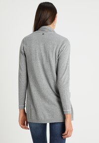 Kvinna med långt brunt hår, iklädd en ljusgrå långärmad cardigan och blå jeans, står med ryggen mot en enfärgad bakgrund.