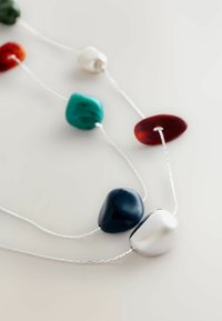 Närbild av ett halsband med oregelbundet polerade stenar i vitt, marinblått, turkos, rött och bärnsten strängda på tunna silverkedjor mot en vit bakgrund.