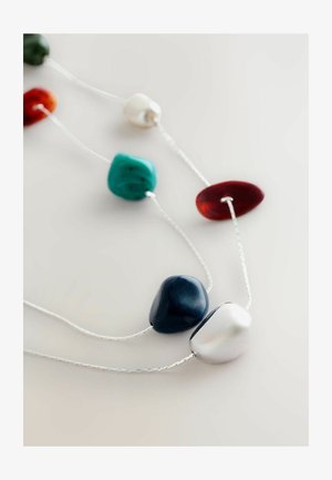 Detailný záber na náhrdelník s nepravidelnými leštenými kameňmi v bielej, námorníckej, tyrkysovej, červenej a jantárovej farbe, zavesenými na tenkých strieborných reťaziach na bielom pozadí.