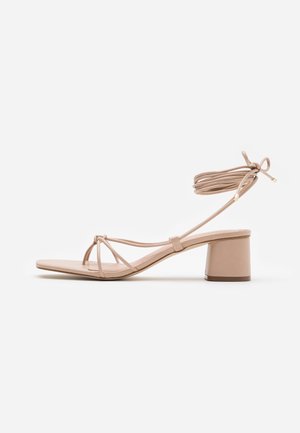 Nude blok-hæl sandal med krydsede remme, ankelbind, flad sål og glat lædertekstur, med et minimalistisk design.
