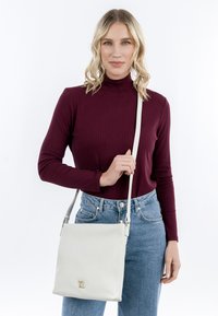Borsa a tracolla bianca in pelle con hardware dorato, forma quadrata e logo impresso. Tracolla regolabile con una superficie liscia.