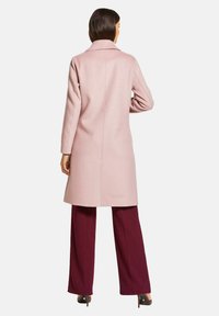 Helle rosa Jacke mit einem Ausschnittkragen, langen Ärmeln und einer strukturierten Oberfläche, kombiniert mit kastanienbraunen weiten Hosen und schwarzenHigh Heels.