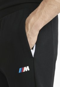 Sorte joggingbukser med tekstureret sidepanel, med en synlig hånd i lommen og et farvet BMW M-logo i rød, blå og hvid.