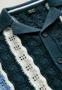 Strickjacke mit Streifen in Marineblau, Weiß und Hellblau, mit einem strukturierten Muster und schwarzen Knöpfen. Etikett zeigt "NEXT, MADE IN CHINA".