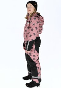 Roze en zwarte sneeuwoverall met sterrenpatronen, elastische manchetten en reflecterende accenten. Draag met een zwarte beanie en donkere sportschoenen.
