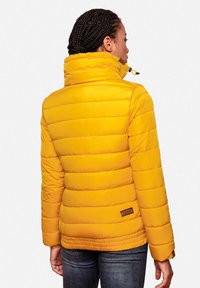 Gele pufferjack met horizontaal gestikte design, hoge kraag en een logo label nabij de zoom. Het materiaal lijkt glanzend en textuur te hebben.