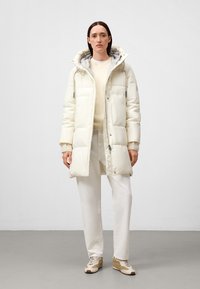 Kvinna klädd i en lång krämfärgad puffjacka med huva, krämfärgad tröja, vita byxor och beiga sneakers, stående på ett grått golv mot en vit bakgrund.