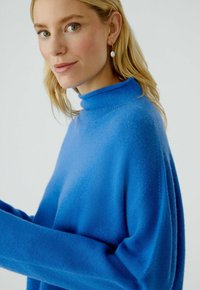 Blauer Rollkragenpullover aus weichem Material mit lockerer Passform, langen Ärmeln und glatter Textur, getragen von einer Person mit blonden Haaren.