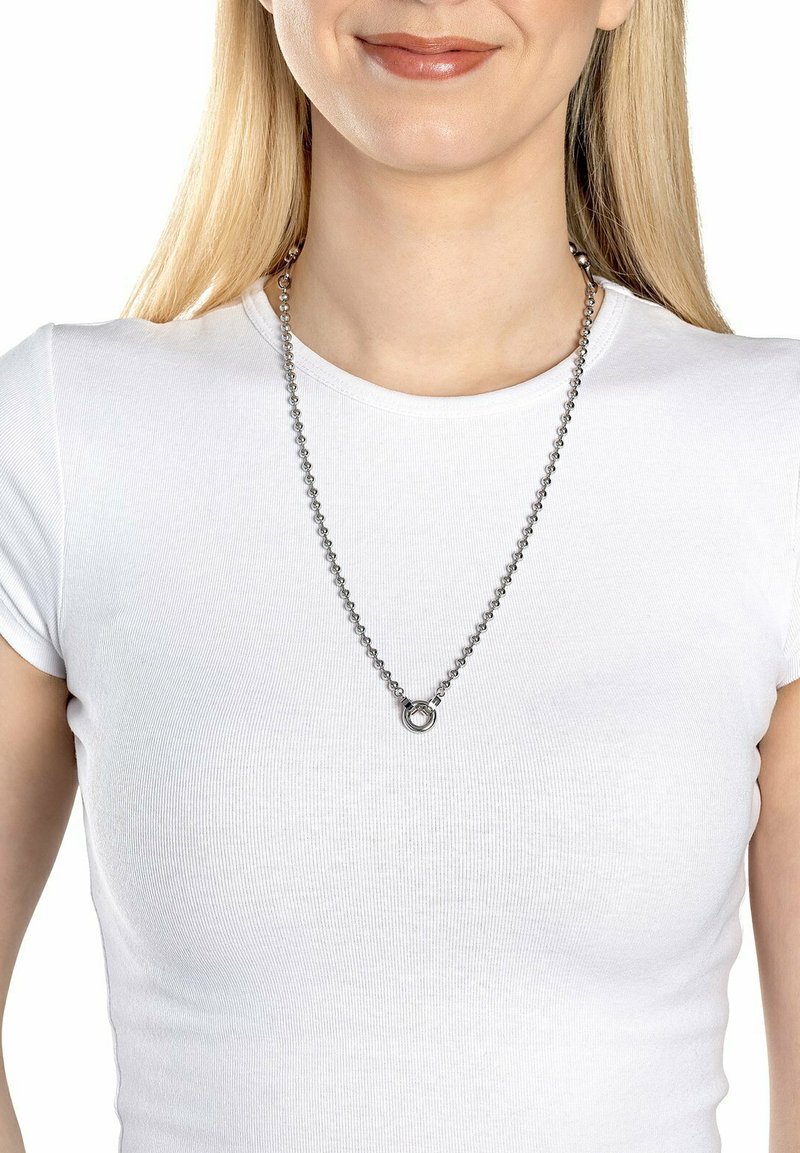Sølv halskæde med en perlekæde og en cirkulær vedhæng, båret over en tætsiddende hvid T-shirt. Kæden har en poleret, reflekterende overflade.