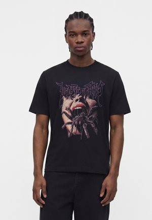 Junger Mann mit geflochtenem Haar, der ein schwarzes T-Shirt mit offenem Mund und großem Spinnengrafik trägt.