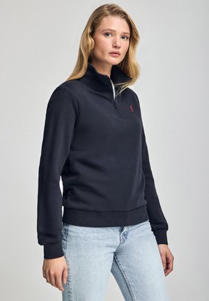Marineblauwe sweatshirt met kwartrits en geribbelde manchetten en zoom, voorzien van een klein rood logo op de borst, gecombineerd met lichtblauwe jeans.