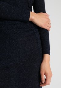 Granatowa sukienka z długim rękawem, wykonana z fakturowanej, błyszczącej tkaniny o dopasowanej sylwetce. Tkanina ma subtelne akcenty brokatowe.