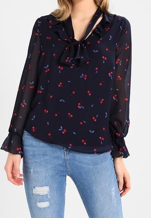 Marineblouse met rood kersen- en blauw bladerenpatroon, ruches aan de manchetten en een strik aan de halslijn, gecombineerd met lichtblauwe gescheurde jeans.