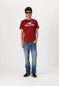 Crvena pamučna majica s logom "HOLLISTER", plave traperice i crne tenisice. Model koji nosi crne sunčane naočale stoji protiv bijele pozadine.