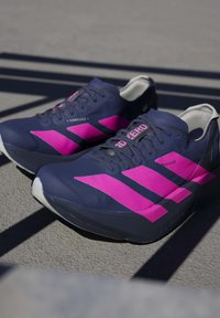 Chaussures de sport bleu foncé avec des bandes roses éclatantes et des lacets noirs reposant sur une surface extérieure texturée avec des ombres.