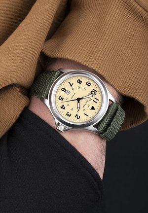 Analog watch with a round stainless steel case, beige dial, large black numbers, and a green woven fabric strap. Features date display.