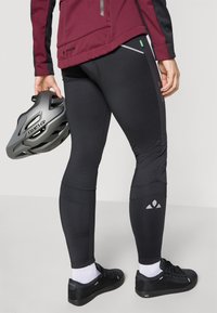 Fekete, rugalmas leggings fényvisszaverő részletekkel, bordó dzsekivel, szürke sisakkal és fekete sportcipővel kombinálva. Jellemzője a feszes illeszkedés és a sima textúra.