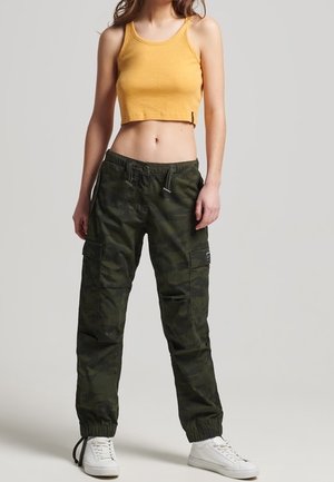 Femme portant un crop top côtelé jaune moutarde, un pantalon cargo camouflage vert foncé et des baskets blanches, debout devant un fond uni.