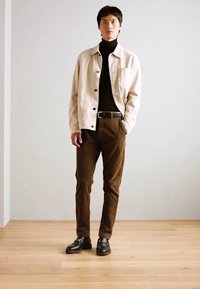 Ljusbeige jacka över en svart polotröja, bruna byxor och svarta skor. Släta material, knapprar och en slim fit-design.