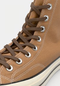 Brune lerret høy-top sneakers med metalløyer, brune lisser, hvite sømmer, og en svart og hvit gummisåle.