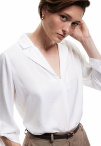 Hvid, letvægtsbluse med V-hals og krave, der har lange ærmer. Bemærkelsesværdig, subtil struktur og let løs pasform. Parret med et brunt bælte.