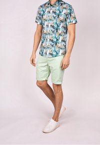 Chemise à manches courtes avec un imprimé tropical dans des tons de vert, orange et blanc. Portée avec un short vert clair et des baskets blanches.