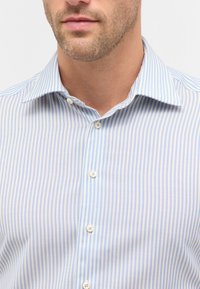 Camicia a righe blu chiaro con colletto standard e bottoni, realizzata in un tessuto liscio. Presenta sottili righe alternate di blu chiaro e blu scuro.